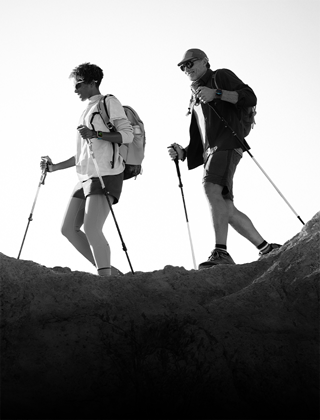 Two people wearing Apple Watch devices and hiking on a rocky trail with poles and gear