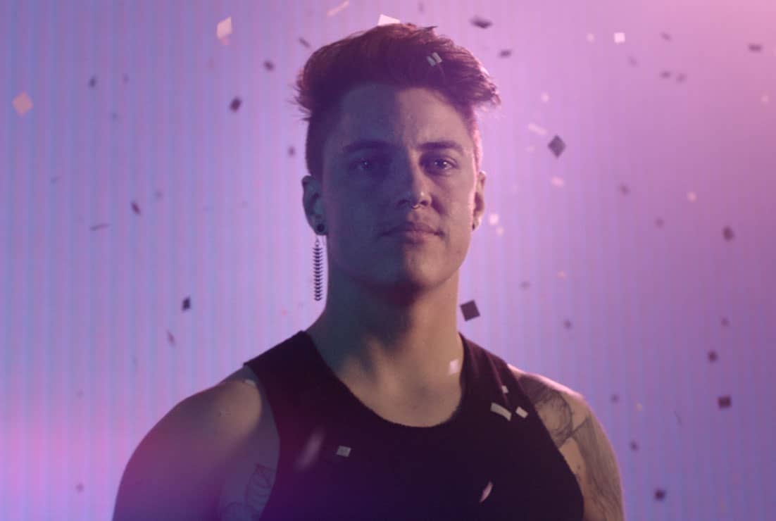 Head shot of non-binary coder, Bode, staring slightly away from camera and standing in front of a pink and purple gradient background with confetti floating behind them.