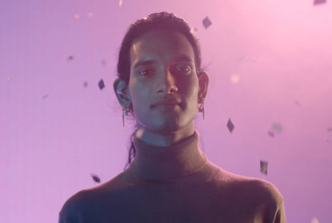Head portrait of Shaneel who is trans non-binary, standing in front of a pink and purple gradient background smiling with confetti floating behind them.