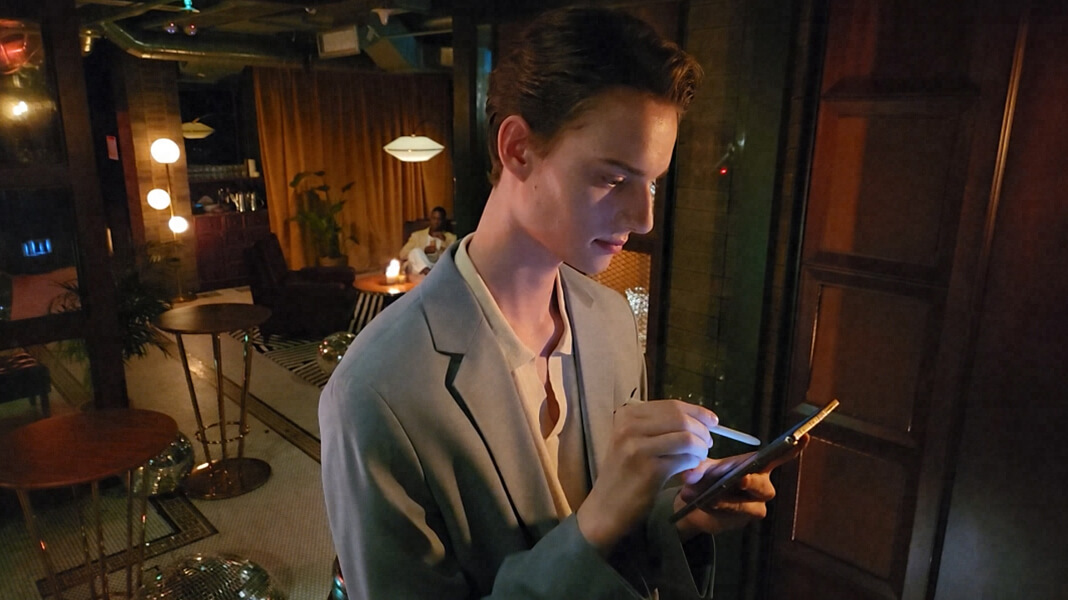 A stylish person in a retro and classy low-lit bar on their Galaxy Z Fold4 using the S pen.