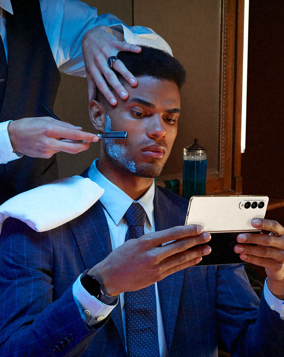 A male model in a suit getting a shave from a barber, using a Galaxy Z Fold4 confidently.