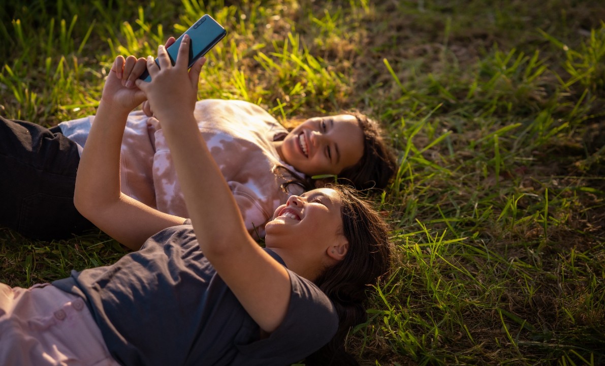 Two kids smiling on their phone while laying on grass